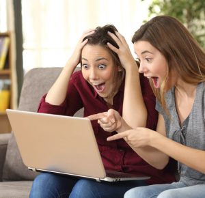 Two astonished roommates watching media content on line with a laptop sitting on a couch in the living room in a house interior